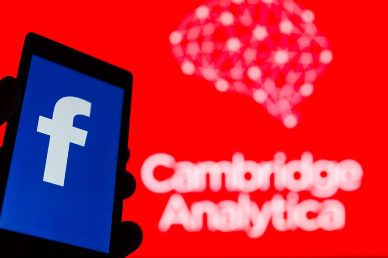 Smartphone in hand with logo of popular social network Facebook. Cambridge Analytica emblem in background.