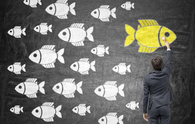 Multiple fish drawn on a chalkboard, with a majority of them being white in color and swimming left and one being yellow and swimming right, used to depict creative differentiation