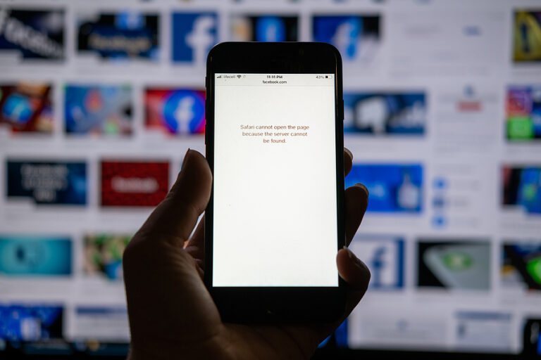 A picture of a person holding a phone that is unable to access Facebook, with a string of Facebook down stories in the background.