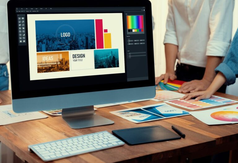 Computer screen displays graphic design program with different design elements like logo and brand colors. Workers gather around a table to brainstorm.