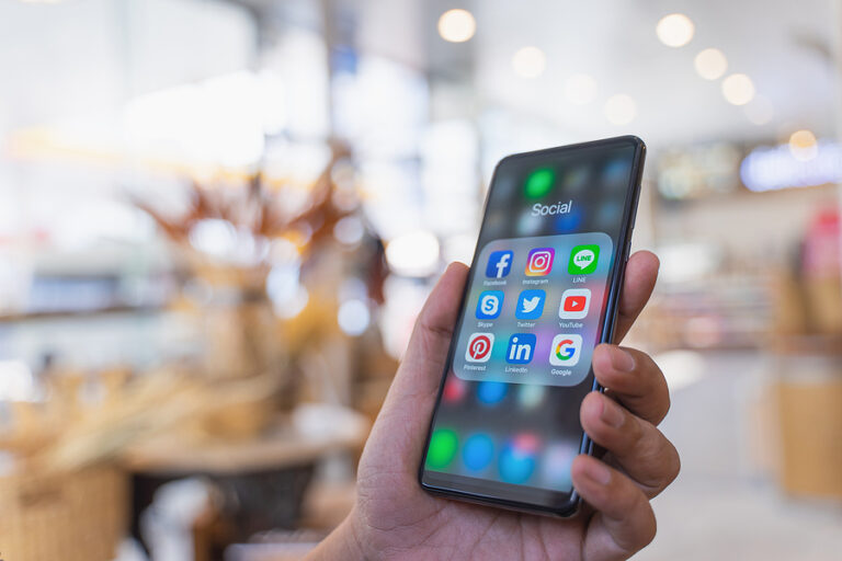 A hand holding a cellphone with social media apps listing on the screen. The man is standing in a store that is blurred out.