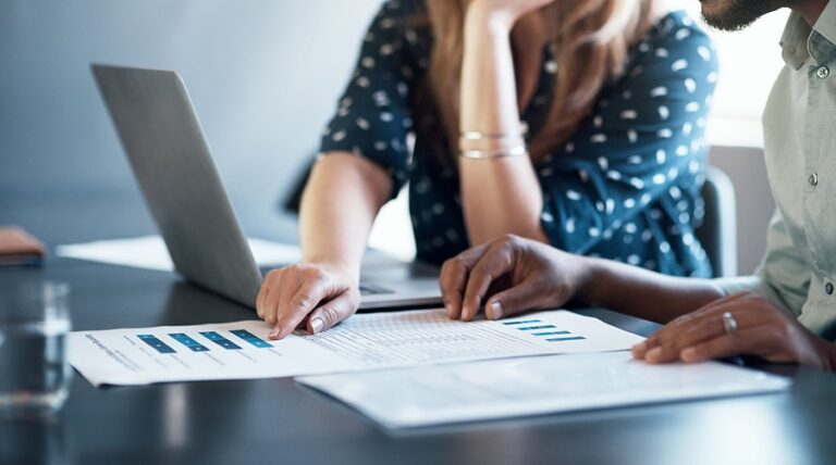 Two women go over papers and laptop with marketing information on them.