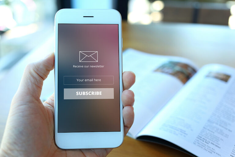 A hand holding a cellphone with a email newsletter subscription form on the screen and a pamphlet atop a desk in the background.