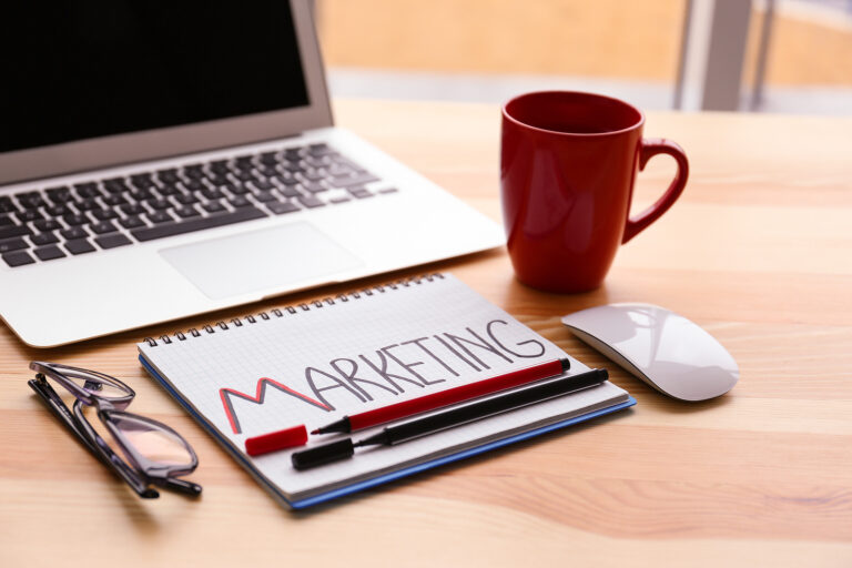 A notebook with "Marketing" written on it next to a pair of glasses, a coffee mug, a computer mouse, and a laptop on top of a desk.