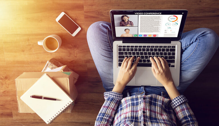 An overhead shot of a person sitting cross-legged with a laptop on their laptop in a video conference. A coffee mug, notepad, box, and cellphone rest on the floor beside her.