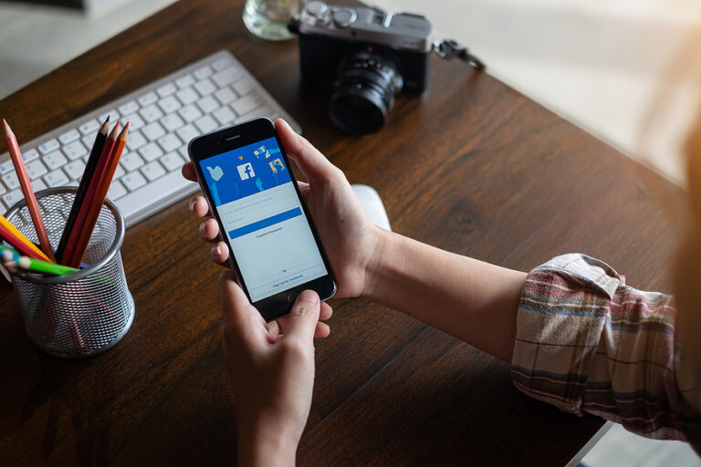 A female photographer using Facebook on her phone, working on a table with a camera, pencil holder, and keyboard on top.