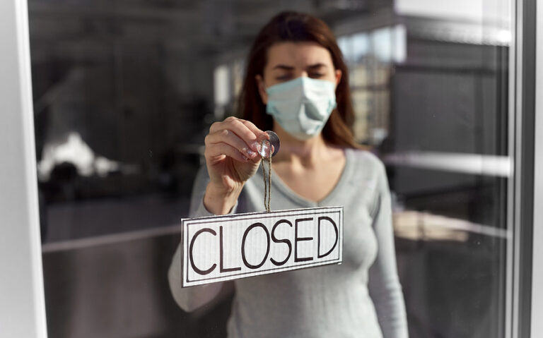 Woman in face mask hangs closed sign on door of shuttered business.