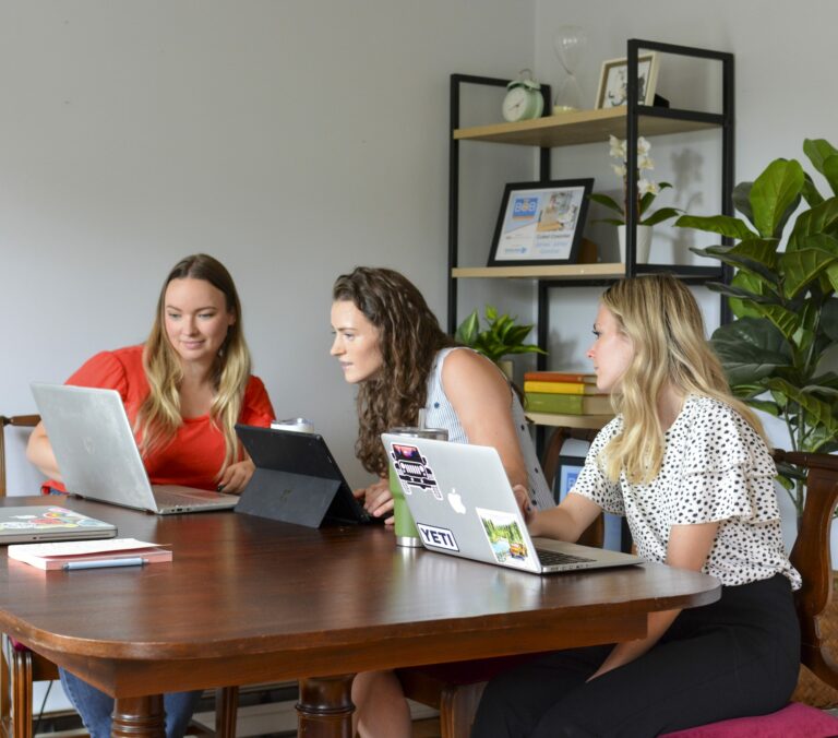 Three ladies sit together at table all working on laptops and looking at something on one person's screen.